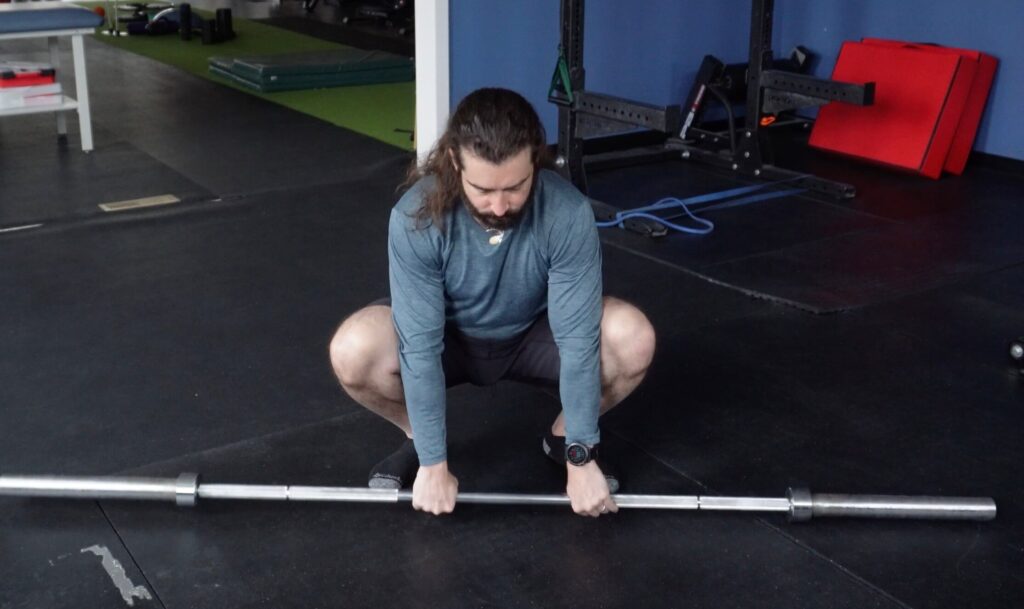 John letting the barbell rest on the floor in front of him while using the weight of the barbell to get himself into a better squat position. This will begin his squat warm up.